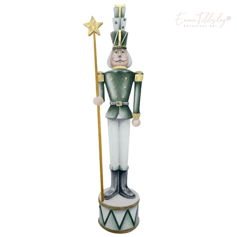 A Christmas nutcracker with gold accents is central to the image. It has shimmering watercolour embellishment, in a traditional soldier uniform.